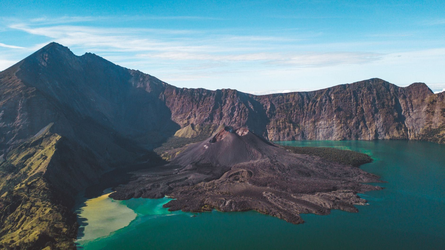 Panoramic view of Mount Rinjani's caldera featuring the turquoise waters of Lake Segara Anak and the Gunung Barujari cone under a blue sky.