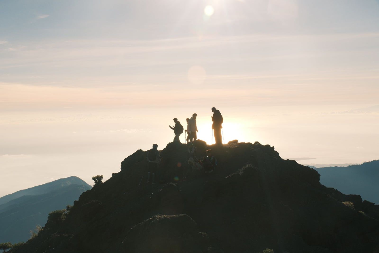 Silhouetted hikers stand on a rocky summit on Mount Rinjani during sunrise.